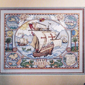 Pode incluir: Um bordado em ponto cruz emoldurado retratando os navios Santa Maria, Pinta e Nina no mar. O design inclui globos, motivos de sol e lua e o texto 1492 COLUMBUS e DISCOVERY OF THE NEW WORLD, em tons de azul, bege e vermelho.