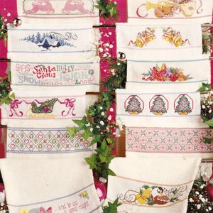 May include: A collection of white fabric towels featuring various cross-stitch holiday designs including reindeer, bells, Christmas trees, a sleigh, holly patterns, and text such as Santa Claus, Ho Ho Ho, and and to all a good night.