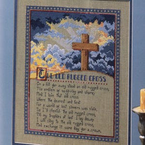 May include: A framed cross-stitch embroidery featuring a wooden cross set against a dark blue and yellow cloudy sky. Below the cross, the text reads The Old Rugged Cross followed by the lyrics of the hymn in dark blue thread on a tan fabric background.