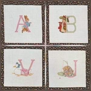 Könnte beinhalten: Vier quadratische Kreuzstich-Paneele mit den Buchstaben A, B, V und U, jeweils mit einer entsprechenden Figur. Die Buchstaben und Figuren sind in Rosa und anderen Farben gehalten, vor einem weißen Hintergrund, umrahmt von einem floralen Rand.