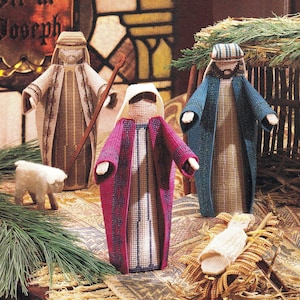 Puede incluir: Un conjunto de nacimiento de lienzo plástico con figuras de José en beige, María en magenta y blanco, y una figura en azul, junto a una pequeña oveja blanca y un bebé en un pesebre. Las figuras presentan texturas y patrones de rejilla.