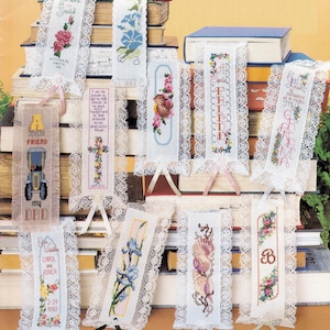 May include: A collection of rectangular fabric bookmarks featuring cross-stitch embroidery designs, including floral patterns, a vintage car, a cross, a bird, seashells, and text. Each bookmark is bordered with white lace and has a small ribbon attached.