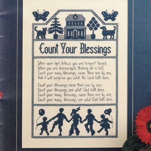 May include: A cross-stitch sampler on cream fabric featuring dark blue thread. The design includes a house, trees, animals, butterflies, and a line of children holding hands. It displays the text Count Your Blessings and a multi-line poem.
