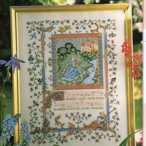 Puede incluir: Obra de arte en punto de cruz enmarcada que representa una escena medieval con una mujer tocando un laúd cerca de un río y un castillo. El marco es dorado. La obra incluye motivos florales y animales, con texto en latín.
