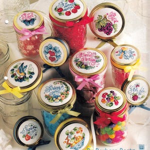 May include: A collection of glass jars with gold metal lids, each topped with a unique cross-stitch design featuring motifs like cherries, grapes, butterflies, and flowers. The jars are decorated with colorful ribbons and filled with various candies.