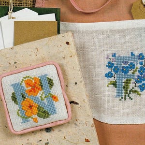 Puede incluir: Un artículo cuadrado bordado a punto de cruz con un borde rosa presenta la letra "N" en azul claro, adornada con flores naranjas y hojas verdes. Otra pieza bordada muestra la letra "T" con flores azules. Suministros de artesanía en el fondo.