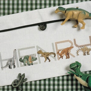 May include: A white fabric pouch with a black snap closure featuring cross-stitched dinosaur motifs and the name HENRY in block letters. A tan plastic dinosaur figure rests on the top edge, and a green plastic dinosaur figure sits below.