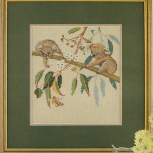 Puede incluir: Obra de arte de punto de cruz de dos koalas en una rama de árbol con hojas verdes, flores blancas y una mariposa. La obra está enmarcada en un marco de madera con un passe-partout verde.