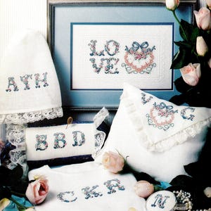May include: A collection of white fabric items featuring floral cross-stitch monograms and heart motifs with lace trim. Included are a framed piece with the word LOVE, a pillow, a small round container, and various linens with letters A, Y, H, S, B, D, C, K, R, and M.