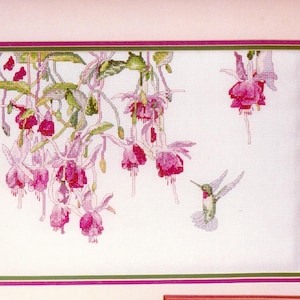 May include: A cross-stitch embroidery piece featuring a cluster of hanging fuchsia flowers in shades of deep pink and purple with green leaves, accompanied by a small hummingbird in flight, all framed by a thin green and magenta border.