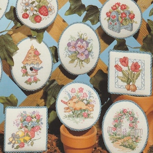 Op de afbeelding: Een verzameling kruissteekontwerpen met bloemen- en tuinthema's. Ontwerpen omvatten aardbeien, viooltjes, tulpen, een vogelhuisje, een konijn en een tuinboog. Elk ontwerp is omlijst met een blauwe, touwachtige rand en geplaatst tegen een blauwe en houtkleurige achtergrond.