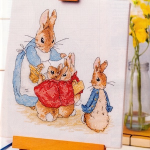 May include: A cross-stitch embroidery on white fabric featuring a group of four brown rabbits. One large rabbit wears a blue coat and white apron, two smaller rabbits wear red jackets, and a fourth rabbit stands to the right in a blue jacket.