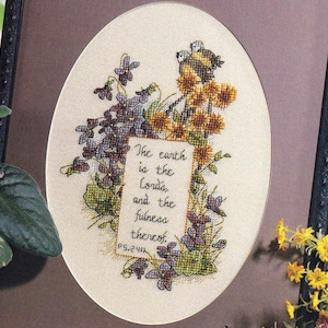 Puede incluir: Un bordado de punto de cruz ovalado en un marco negro con violetas moradas y flores amarillas que rodean un bloque de texto rectangular central que dice: The earth is the Lord's, and the fulness thereof. Ps. 24:1.