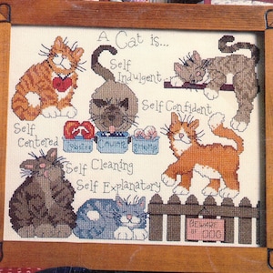 May include: A framed cross-stitch sampler featuring various cats and the text A Cat is... Self Indulgent, Self Confident, Self Centered, Self Cleaning, Self Explanatory, Lobster, Caviar, Shrimp, and Beware of Dog. The wooden frame has heart-shaped corner accents.