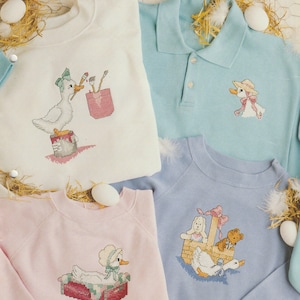 Può includere: Una collezione di quattro felpe e una polo color pastello con ricami a punto croce raffiguranti anatre. I capi sono color crema, azzurro e rosa pallido, decorati con motivi dettagliati di anatre in varie scene.