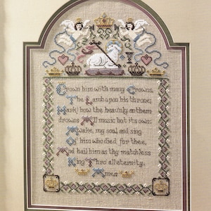 May include: A framed cross-stitch sampler on beige linen featuring a white lamb, two angels, and multiple crowns. The text reads: Crown him with many Crowns, The Lamb upon his throne; Hark! how the heavnly anthem drowns All music but its own; Awake, my soul, and sing Of him who died for thee, And hail him as thy matchless King thro' all eternity. Amen.