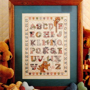 May include: Framed cross-stitch alphabet sampler with a teddy bear theme. The letters are in various patterns and colors, with a border of hearts and the alphabet. The frame is wood, and the background is a light blue.