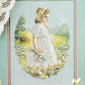 May include: A cross-stitch embroidery depicting a child with a blonde bonnet and a white dress standing in a field of colorful wildflowers. The scene is framed by a green border with a thin red line, set against a light blue background.