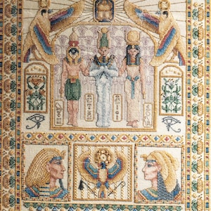 Peut inclure: Une broderie au point de croix détaillée représentant des motifs égyptiens anciens, incluant trois personnages centraux, des divinités ailées, des hiéroglyphes et deux portraits de pharaons, encadrés par une bordure dorée, bleue et verte.