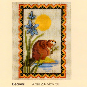 May include: Cross-stitch design featuring a brown beaver, blue flower, and yellow sun. The image is framed with a decorative border in black, orange, and green. Text at the bottom reads "Beaver April 20-May 20".