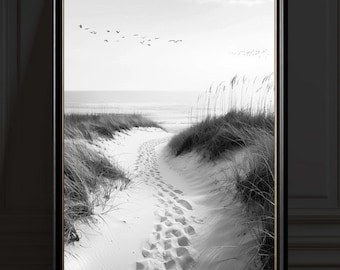 Puede incluir: Impresión en blanco y negro enmarcada de una escena de playa. Un camino arenoso con huellas conduce al océano, flanqueado por dunas con hierba alta. Aves volando en el cielo. El marco es negro.