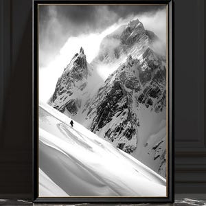 May include: Monochrome framed print depicting a snow-covered mountain range. A skier descends the slope. The artwork is encased in a black frame with a gold inner edge. The mountain peaks are partly hidden by clouds.