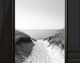 Puede incluir: Impresión en blanco y negro enmarcada de un camino arenoso que conduce al océano. El camino está flanqueado por dunas con hierba alta. El cielo es brillante y el agua está tranquila. La obra de arte se muestra en un marco negro.