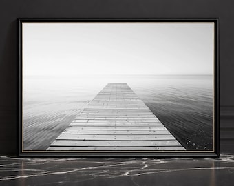 Puede incluir: Impresión enmarcada en blanco y negro de un muelle de madera que se extiende hacia aguas tranquilas. El muelle está hecho de tablas de madera de color claro, y el agua y el cielo se mezclan en una escena monocromática suave. El marco es negro.