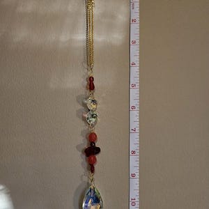 May include: A decorative hanging crystal ornament, approximately 25 cm long, with a gold chain and various red and clear crystal beads. The bottom features a large, iridescent teardrop-shaped crystal.