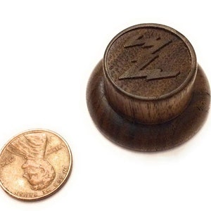 May include: A dark brown wooden knob with a raised, stylized lightning bolt design. The knob is approximately the size of a UK penny.