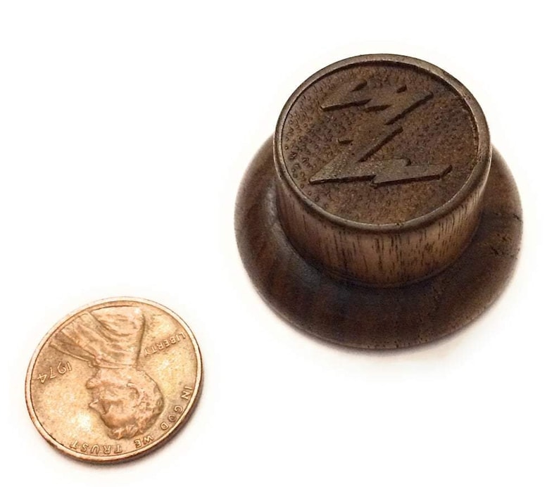 May include: A dark brown wooden knob with a raised, stylized lightning bolt design. The knob is approximately the size of a UK penny.