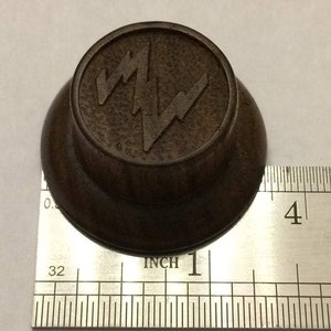 May include: A dark brown wooden knob with a raised, stylised lightning bolt design. The knob is approximately 1 inch in diameter.