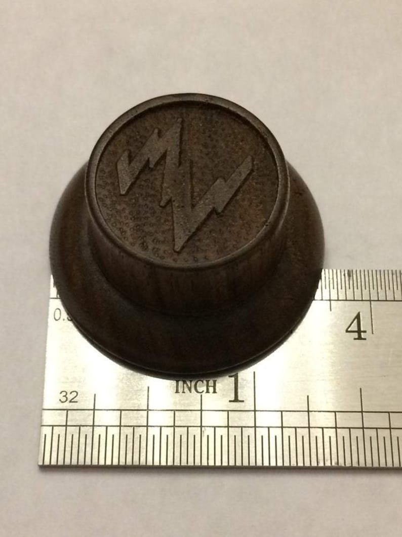 May include: A dark brown wooden knob with a raised, stylised lightning bolt design. The knob is approximately 1 inch in diameter.