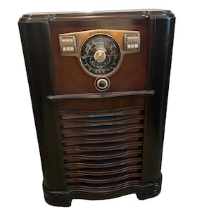 May include: An antique Zenith radio with a dark wood cabinet. The radio features a round dial with a brass-colored face and the Zenith logo. The front has a series of horizontal slats for the speaker.