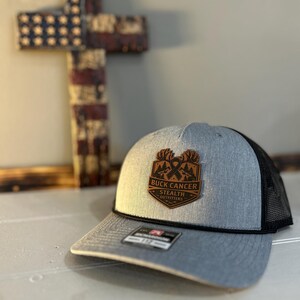 May include: A gray and black trucker hat with a patch that reads "BUCK CANCER STEALTH OUTFITTERS". The hat has a curved brim and a black mesh back. A wooden cross with an American flag design is in the background.