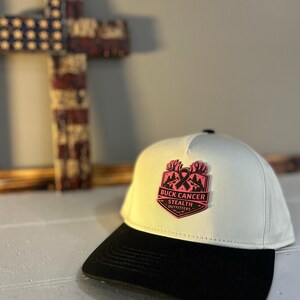 May include: A white baseball cap with a black brim. The cap features a pink and black patch with the text "BUCK CANCER STEALTH OUTFITTERS" and a pink ribbon and mountain design.