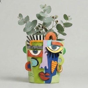 Könnte beinhalten: Eine farbenfrohe, abstrakte, gesichtsformige Keramikvase mit grünen, blauen, orangefarbenen und schwarzen Akzenten. Die Vase enthält grüne, belaubte Zweige. Die Website-Adresse www.LeVeridianStudio.etsy.com befindet sich am unteren Rand des Bildes.