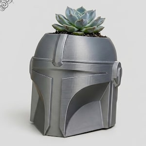 May include: A silver Mandalorian helmet-shaped planter with a succulent plant. The planter has a textured surface and is designed to resemble the iconic helmet from the Star Wars series. The succulent has blue-green leaves. The website address www.LeVeridianStudio.etsy.com is at the bottom.