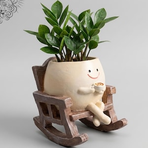 Op de afbeelding: Een grillige crèmekleurige plantenbak in de vorm van een ei, met een lachend gezicht, een kopje koffie vasthoudend en zittend in een bruine schommelstoel. De plantenbak is gevuld met groene bladplanten. De website www.LeVeridianStudio.etsy.com staat onderaan.