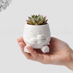 May include: A white ceramic planter shaped like a rounded character with a smiling face, holding a small white cat figure on its front. The planter contains a green and reddish-tipped succulent plant and features two small feet at the base.