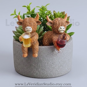 May include: A grey cylindrical planter filled with green succulents, featuring two brown Highland cow figurines perched on the rim. One cow holds a miniature mug of beer, while the other holds a small glass of red wine. Text reads LeVeridianStudio.etsy.com.
