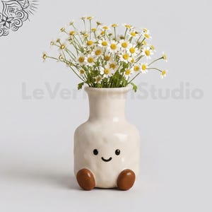 May include: A cream-coloured ceramic vase with a textured surface, featuring a simple black painted face with two eyes and a smile. Two brown rounded feet are attached at the base. The vase holds a bouquet of white daisies with yellow centres.