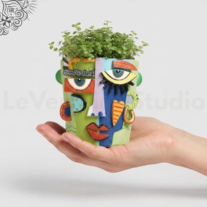 Può includere: Un vaso in ceramica multicolore a forma di volto con tratti astratti, tra cui una base verde e blu, un naso centrale e occhi stilizzati. Il vaso è riempito con piccolo fogliame verde e presenta un design testurizzato dipinto a mano.
