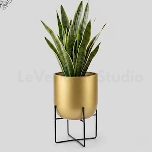 Op de afbeelding: Een sansevieria met gevlekte groen-gele zwaardvormige bladeren in een gladde, goudkleurige cilindrische plantenbak. De bak rust op een dunne, zwarte metalen draadstandaard met een kruisvormige voet. De tekst luidt LeVeridian Studio.