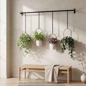 May include: A black metal wall-mounted rod with four hanging plant holders, each containing a white pot with a green plant. The rod is attached to the wall above a light-colored wooden bench with a woven seat and a draped fabric cloth.