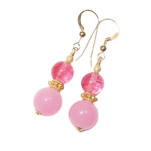May include: A pair of dangle earrings featuring pink beads and gold-colored accents. The earrings have a gold-colored hook and are composed of two pink beads, one round and one textured, separated by gold-colored decorative elements.