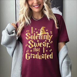 May include: Burgundy t-shirt with gold text that reads "I Solemnly Swear that I Graduated." The design includes a wizard hat and stars. The shirt is short-sleeved.