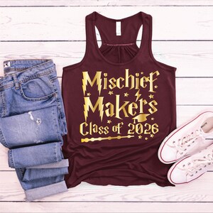 May include: A maroon tank top with gold text that reads "Mischief Makers Class of 2026". The tank top is paired with blue jeans and white sneakers on a white wooden surface.