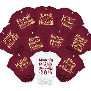 May include: A selection of burgundy and white t-shirts with gold lettering. The shirts display phrases such as "Mischief Mastermind," "We Are Up to No Good," and "Mastering Mischief Since 2011." Perfect for themed gatherings or groups.