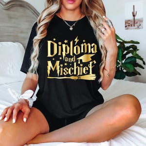 May include: Black t-shirt featuring the gold text "Diploma and Mischief". The design incorporates a lightning bolt, stars, a mortarboard, and a broom. The short-sleeved shirt is likely made from a soft fabric.